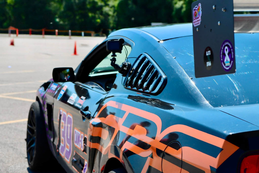 A black race car with orange and purple graphics, featuring a rear-mounted camera and a black vertical panel with stickers, photographed at an autocross